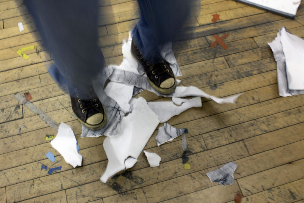 Pair of feet wearing sneakers surrounded by torn drawings on wood floor.