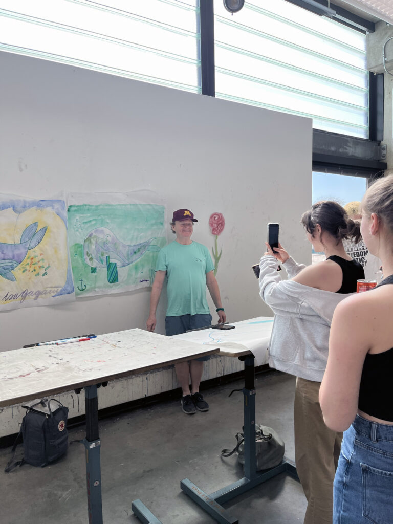 Student takes a photo of another student, standing behind folding table and in front of artworks hung on wall.