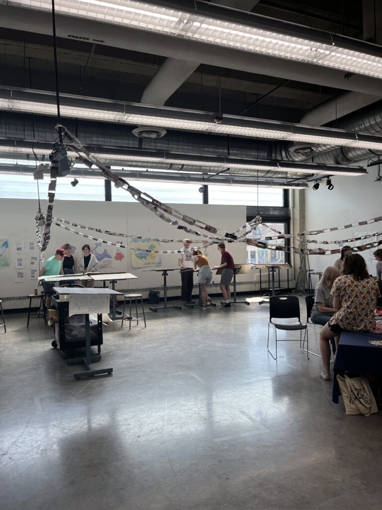 Classroom with streamers hung, folding tables and chairs, and students seated and standing.