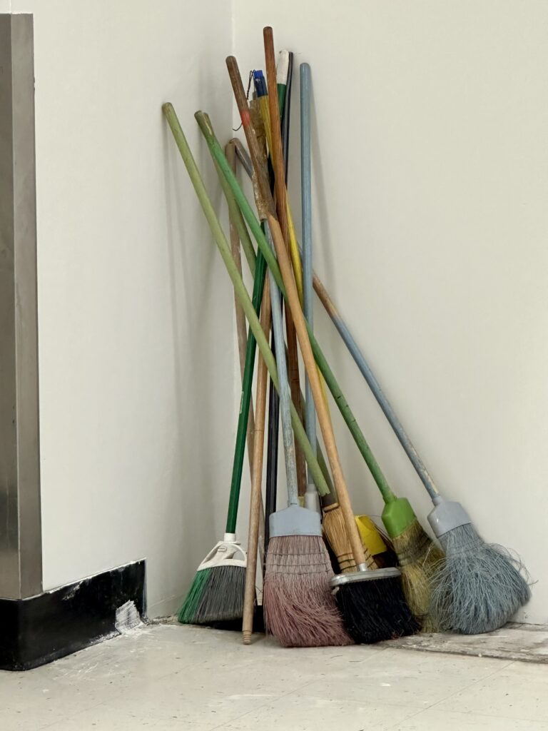 Stack of brooms in various colors leans against corner of white wall.