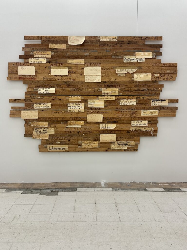 Artwork made from strips of wood labeled with names of colors hangs on white wall in front of linoleum floor.