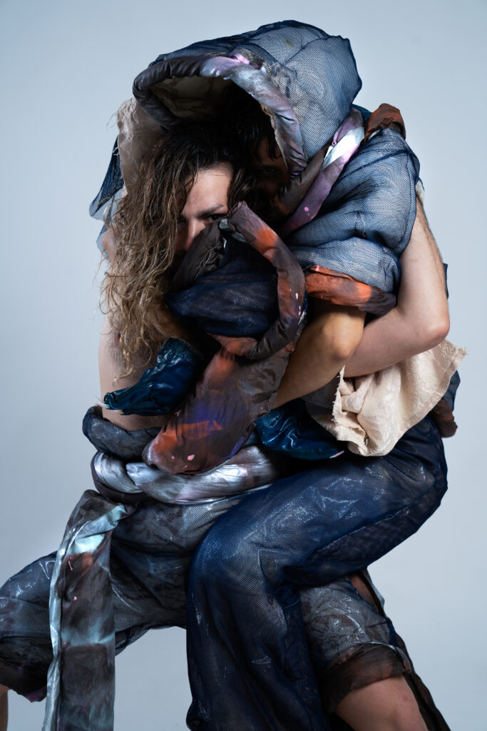 Two dancers embrace each other wrapped in blue fabric, with person with long hair peeking out at the camera.