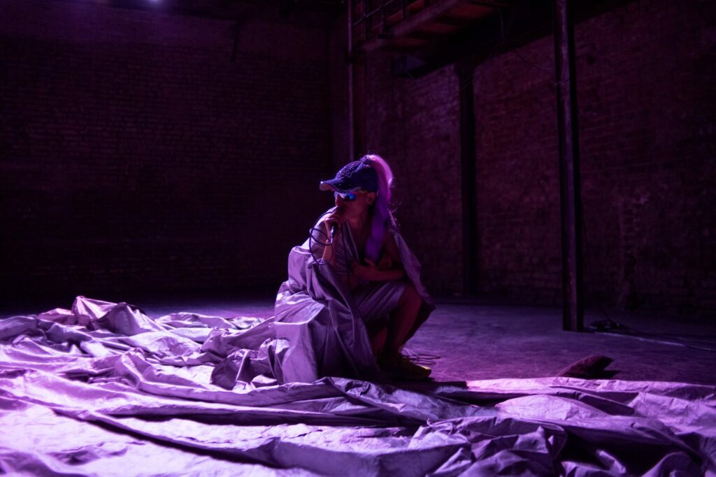 Performer wearing baseball cap wraps herself in tarp and holds microphone to mouth, bathed in purple light.