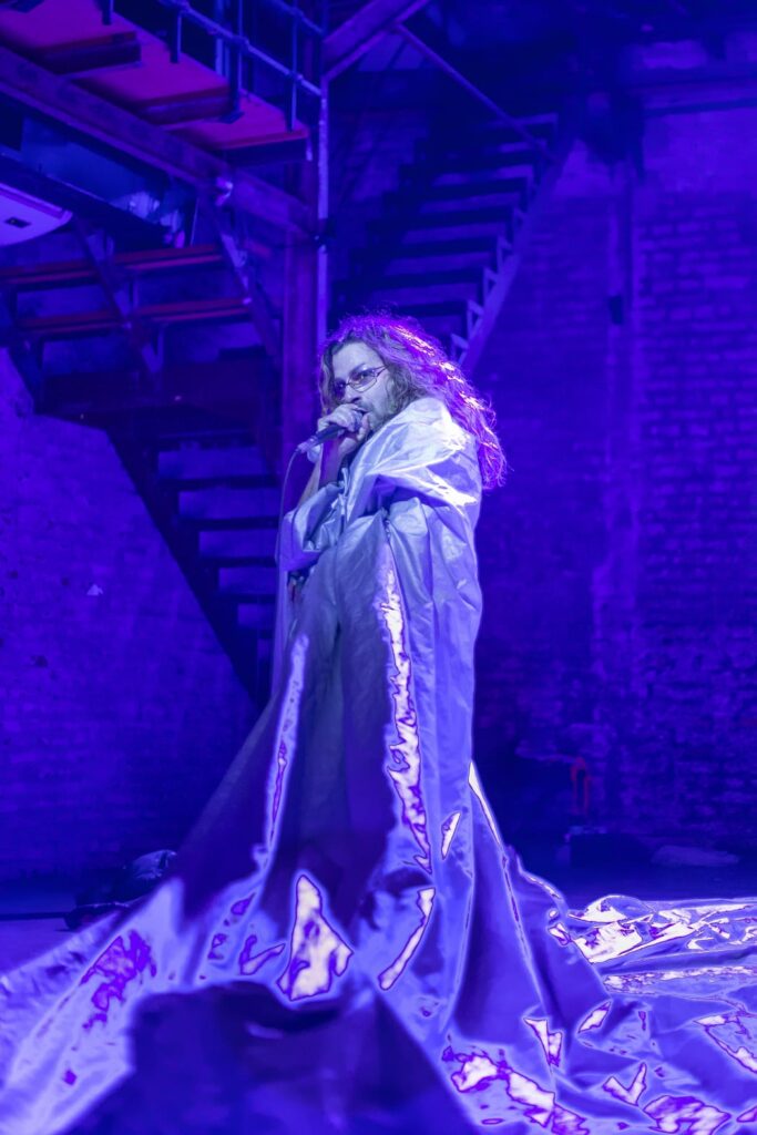 Performer with long hair wraps themselves in fabric and holds microphone to mouth, bathed in blue light.