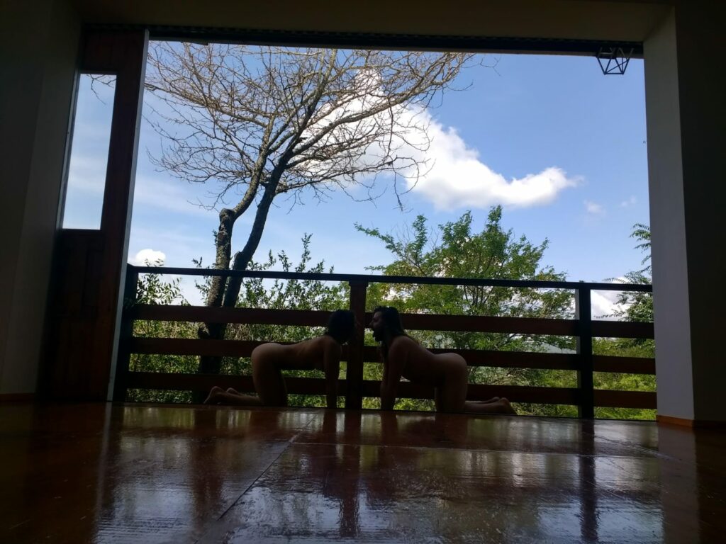 Two dancers kneel on all fours facing each other in open air studio, with trees and blue sky in background.