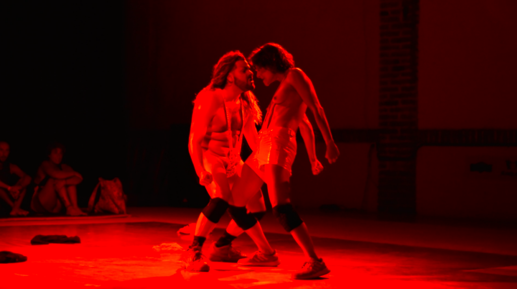 Two performers look each other in the eye and step toward each other, bathed in red light.