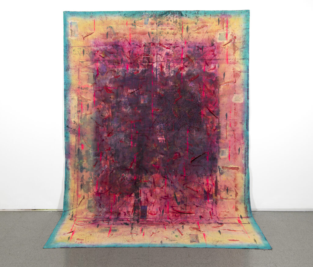 Painted canvas with dark red, pink, orange, and teal colors emanating from center, hung on white wall and draped onto floor.