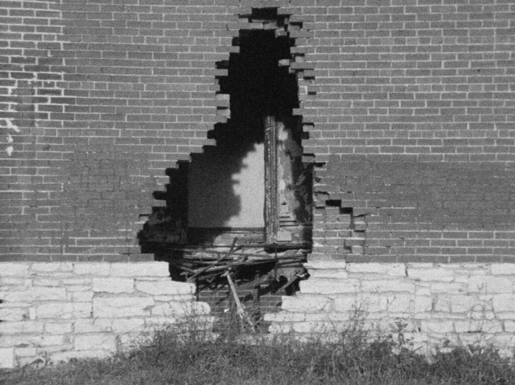Black and white film still of brick building with hole in wall and long grass.