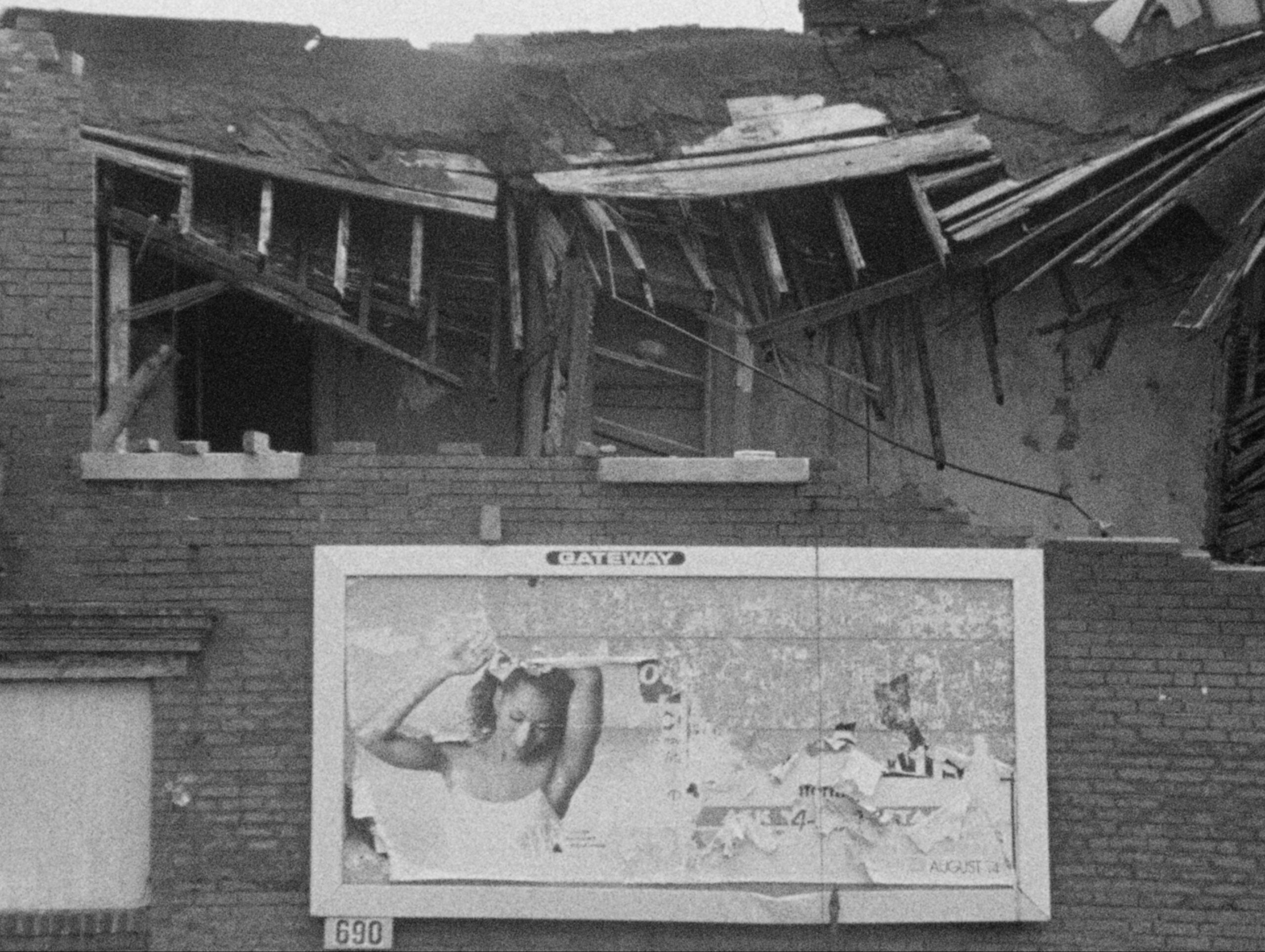 Black and white film still of brick building with roof caving in and tattered billboard.