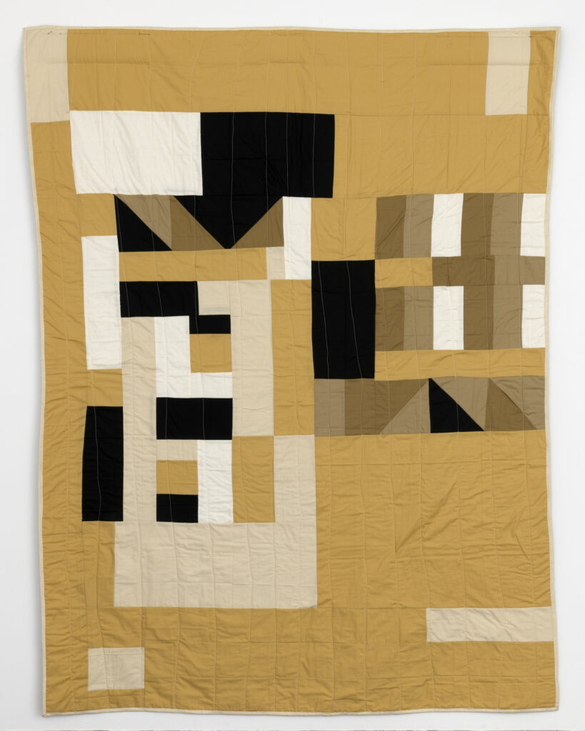Quilt with shades of tan, black, and brown in asymmetrical pattern.