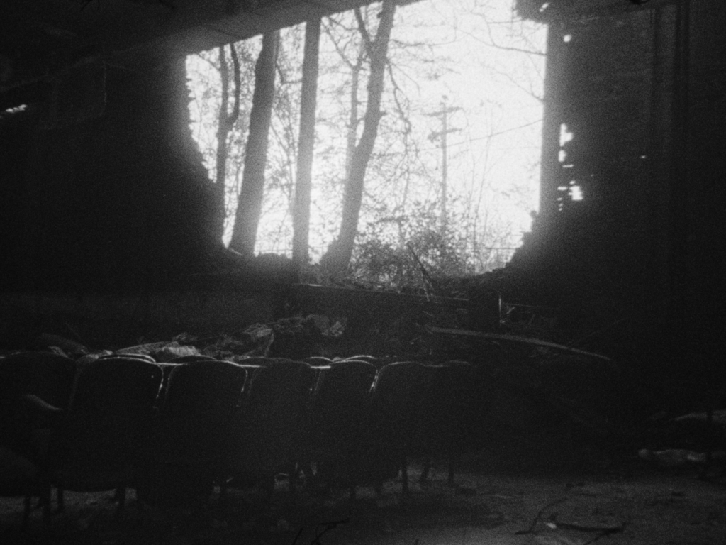 Black and white film still of building interior with brick wall open to sky and trees.