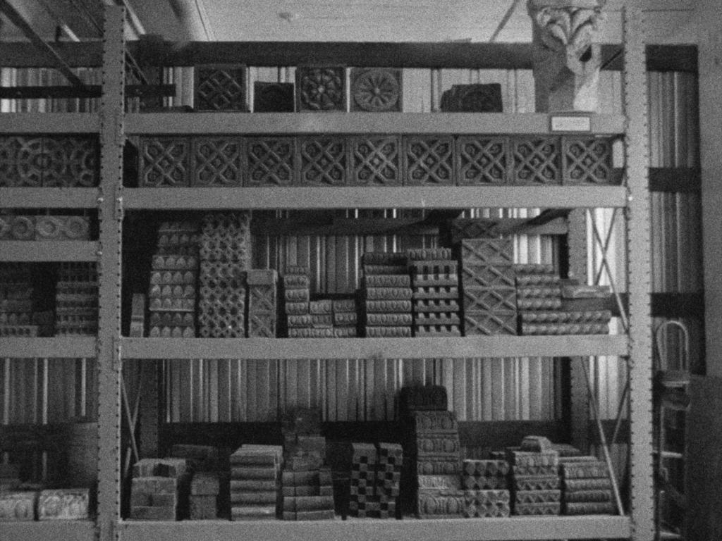Black and white film still: rack of shelves with stacks of patterned bricks.