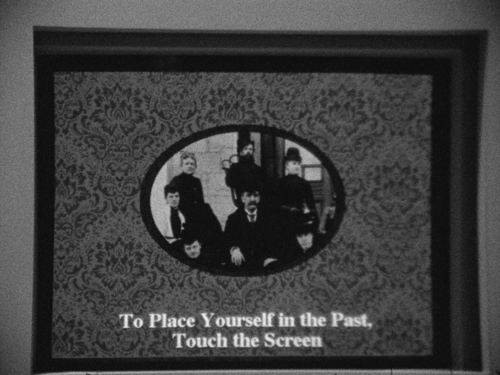Black and white film still of vintage picture frame with group portrait and subtitle reading: "To Place Yourself in the Past, Touch the Screen".