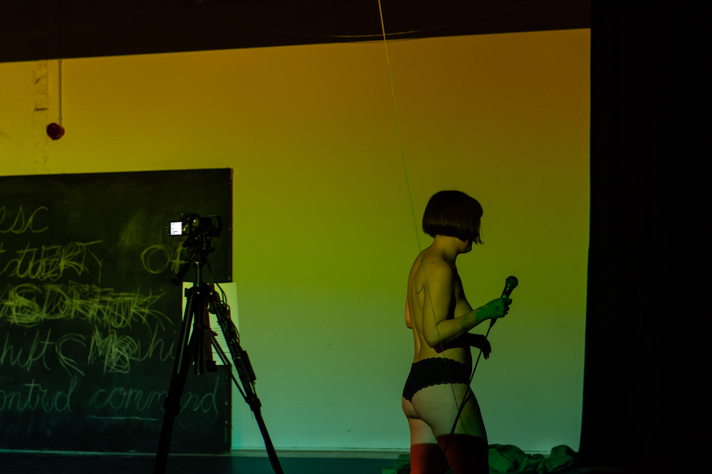 White woman in black panties carries a microphone away from the camera, with video camera on tripod, chalkboard, and blue-green light behind her.