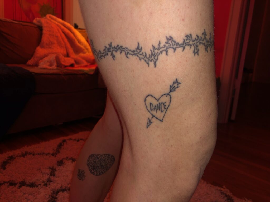 Leg of a person with light skin, tattoo surrounding the thigh and a tattoo of a heart with an arrow through it and the word DANCE inside, above the knee.
