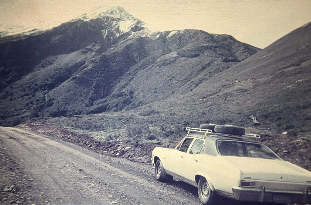 Old photograph of white car on gravel road with mountains in background