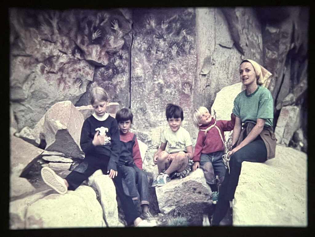 Four small children and their mother sitting on rocks with a rock wall with painted hands behind them