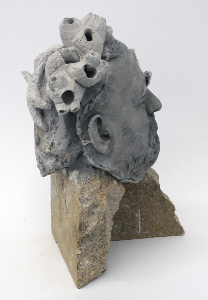 Face with closed eyes and beard with barnacles on head cast in concrete on cement stand