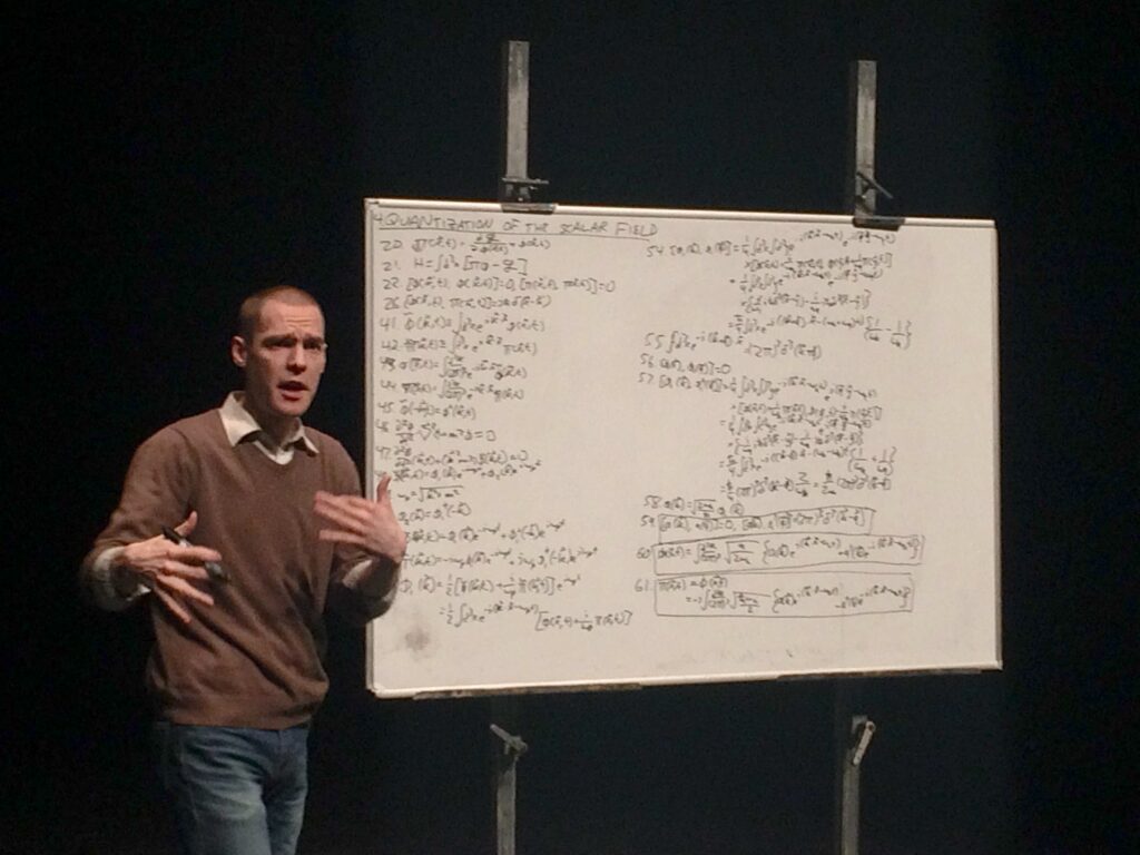 Person stands in front of white board with many equations mid speach