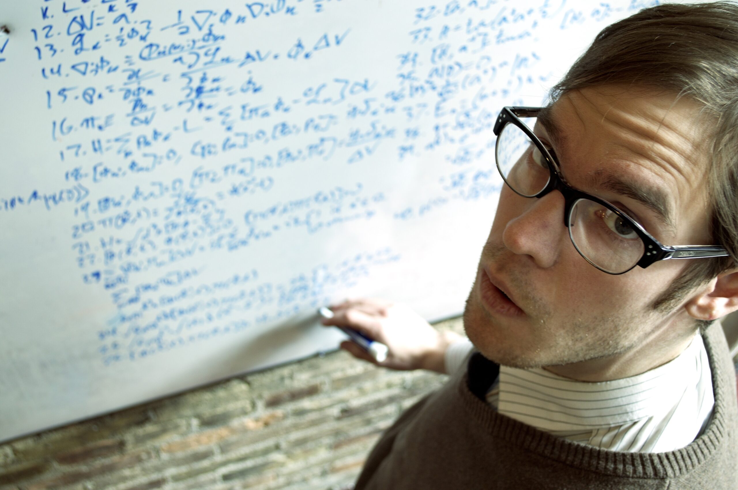 Person with glasses staring up at camera, writing equations on whiteboard in background