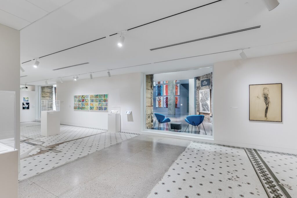 White gallery space with tile floors and artwork hung on walls, and a seating areas with two blue chairs