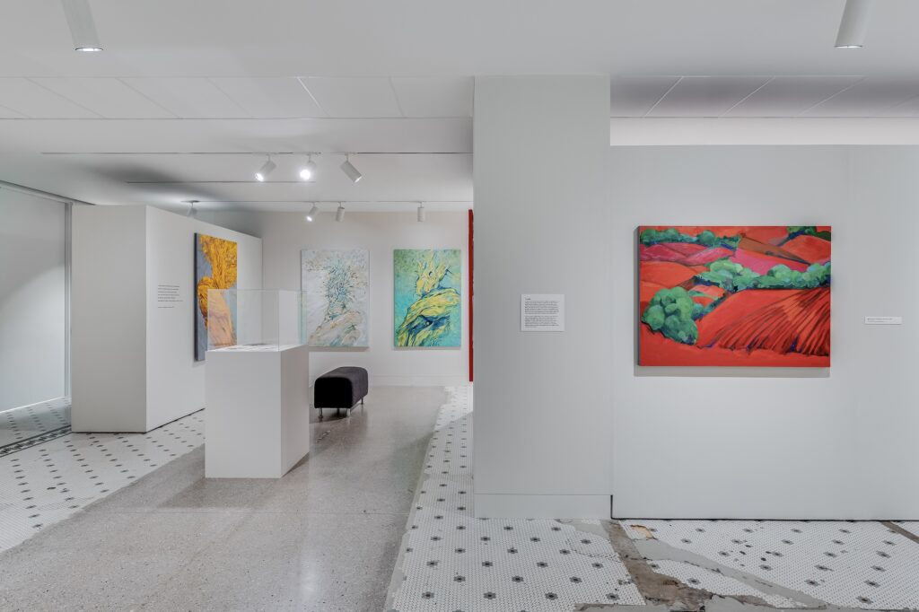 White gallery space with painting of red landscape in foreground, podium, bench, and colorful tree images in background