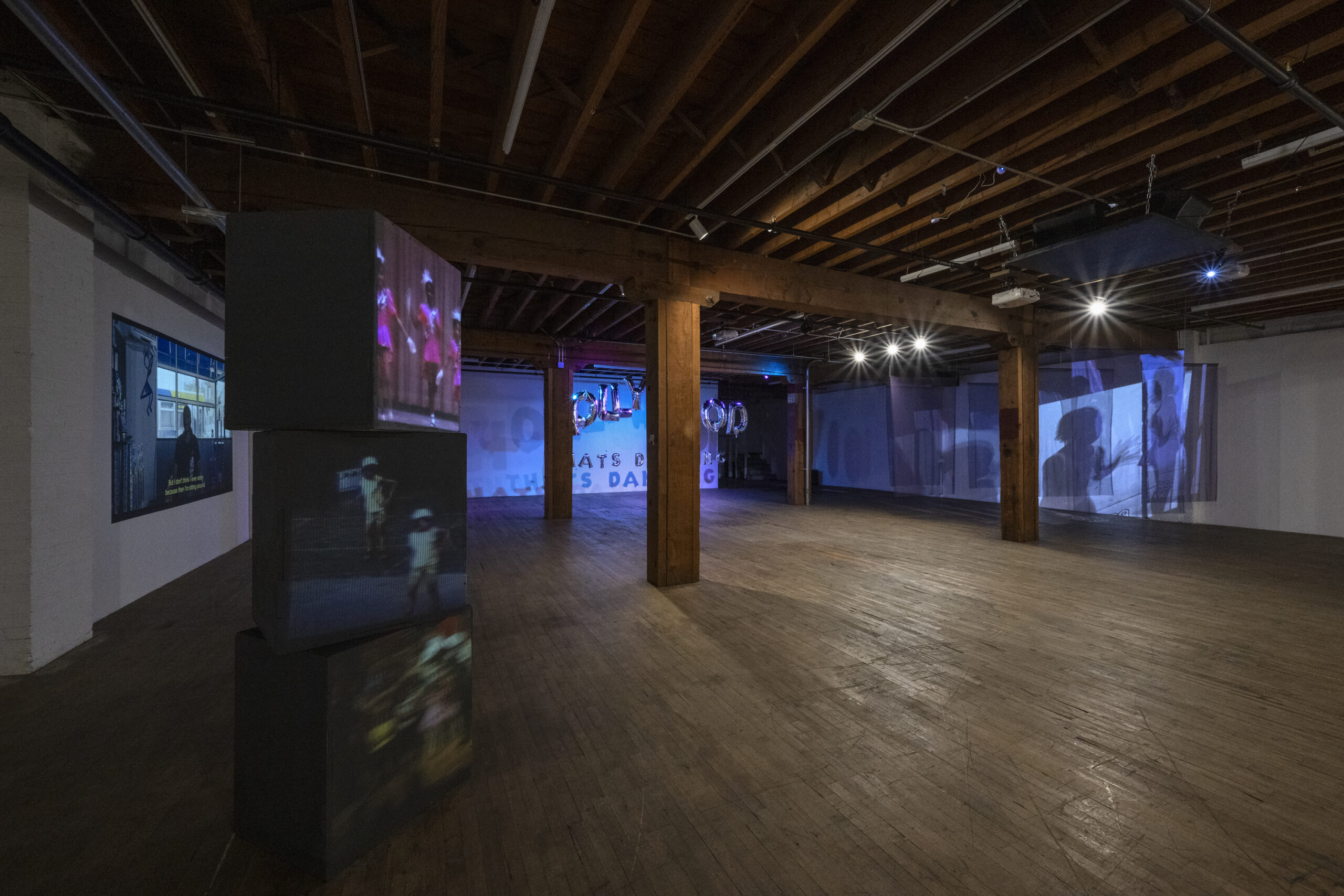 View of a multimedia art exhibition in a dimly lit space with exposed wood beams