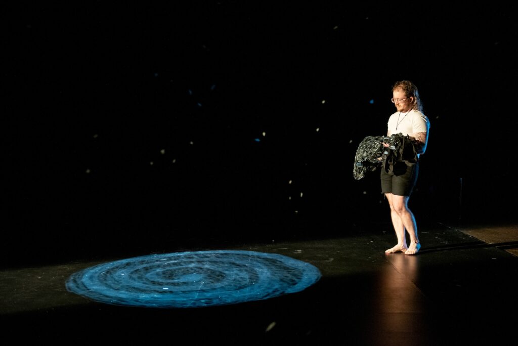 Person stands on black stage carrying fabric towards dark swirl projected on floor.