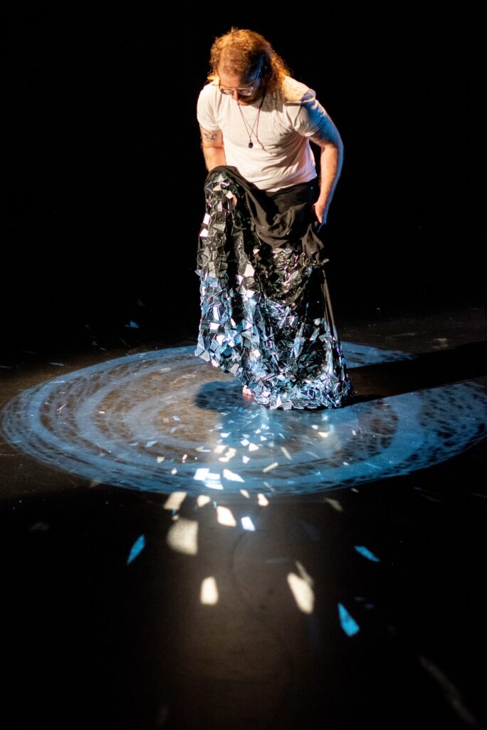 Person stands on dark swirl projected on black floor, holding cloth with mirror shards reflecting light.