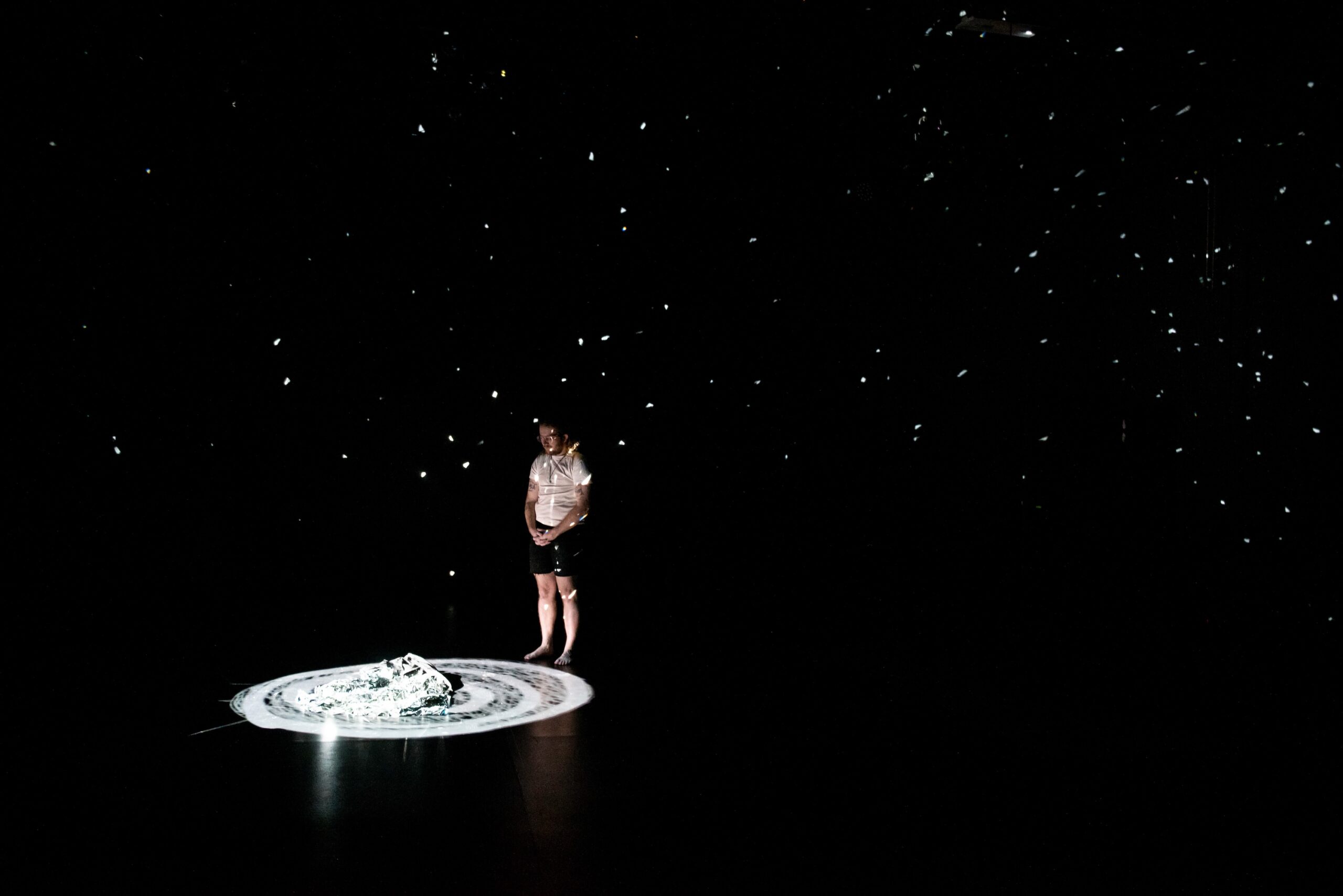 Person stands amidst darkness and small spots of light, staring at cloth in a swirl of white light on the floor.