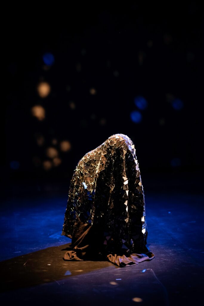 Figure of person, shrouded in dark cloth with mirror shards reflecting light on dark stage