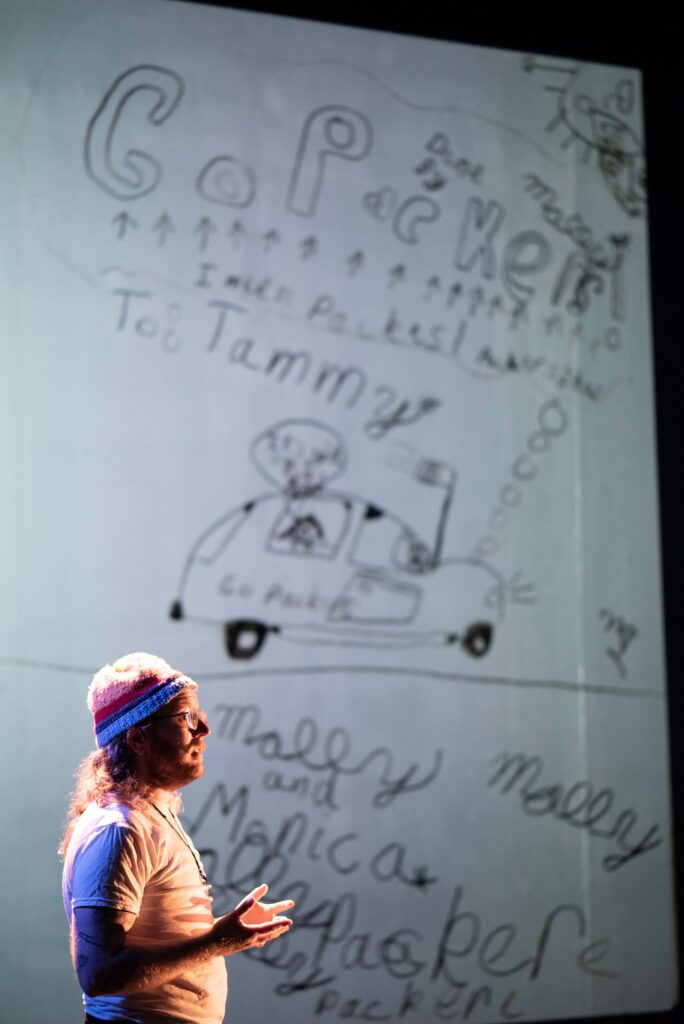 Person stands in front of screen showing a young person's writing and drawings, with mouth open and hands gesturing by waist.