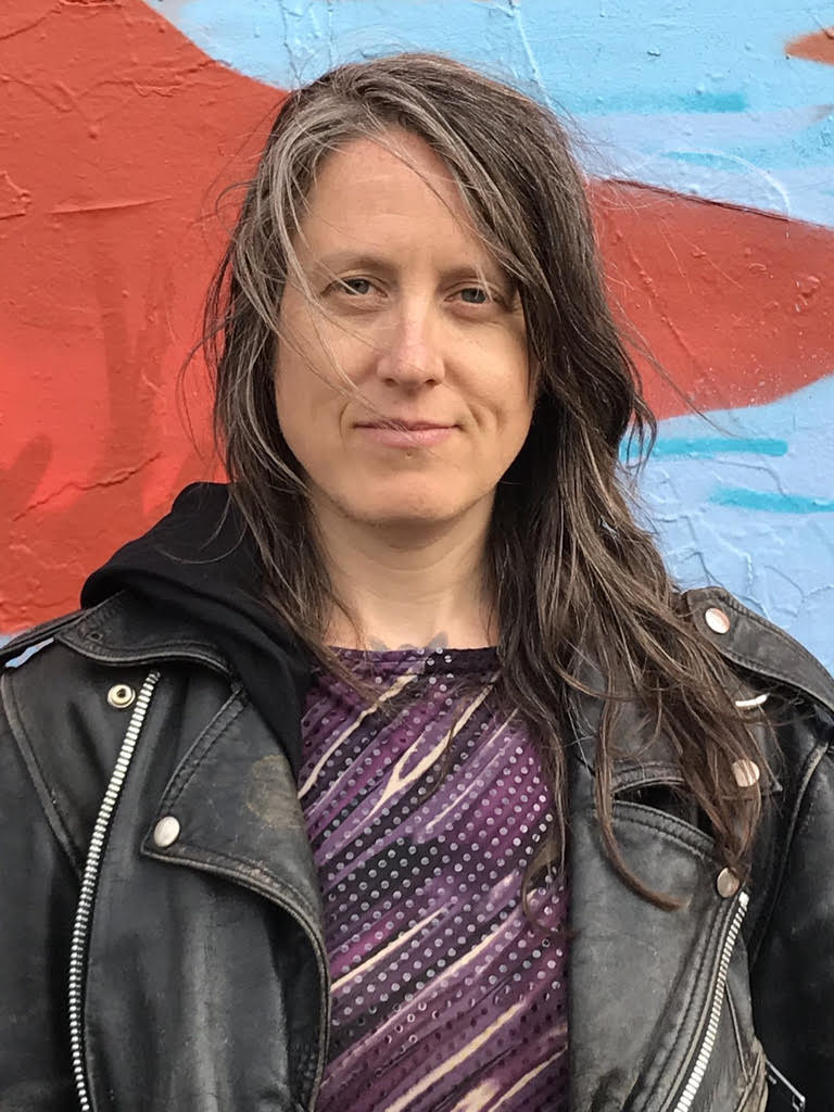 Woman with brown greying hair in leather jacket with slight smile looks at the camera.