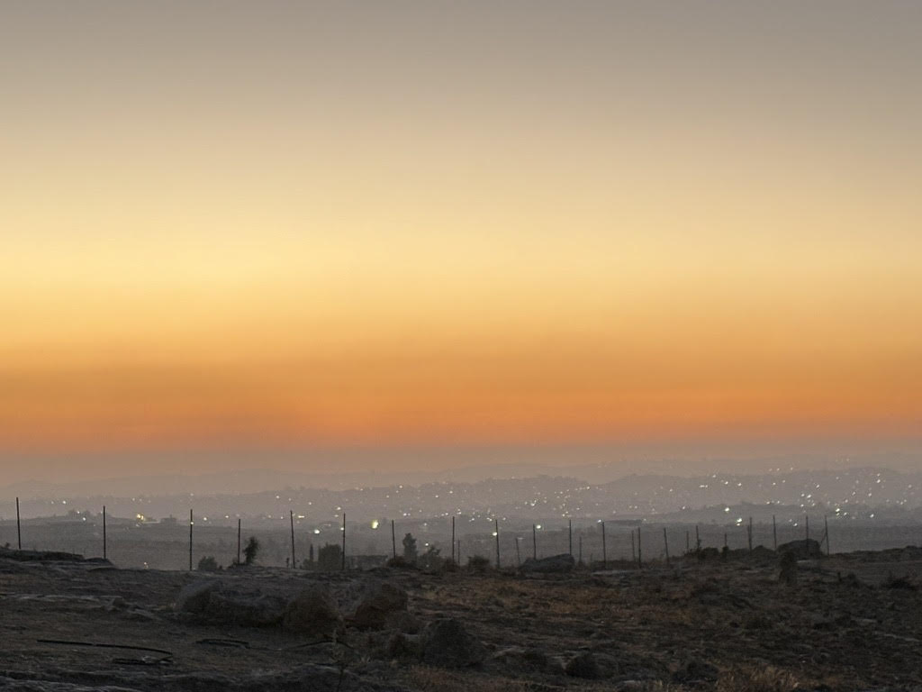 Hazy orange sunset overlooking a barren hillside and lights from a town in the distance.