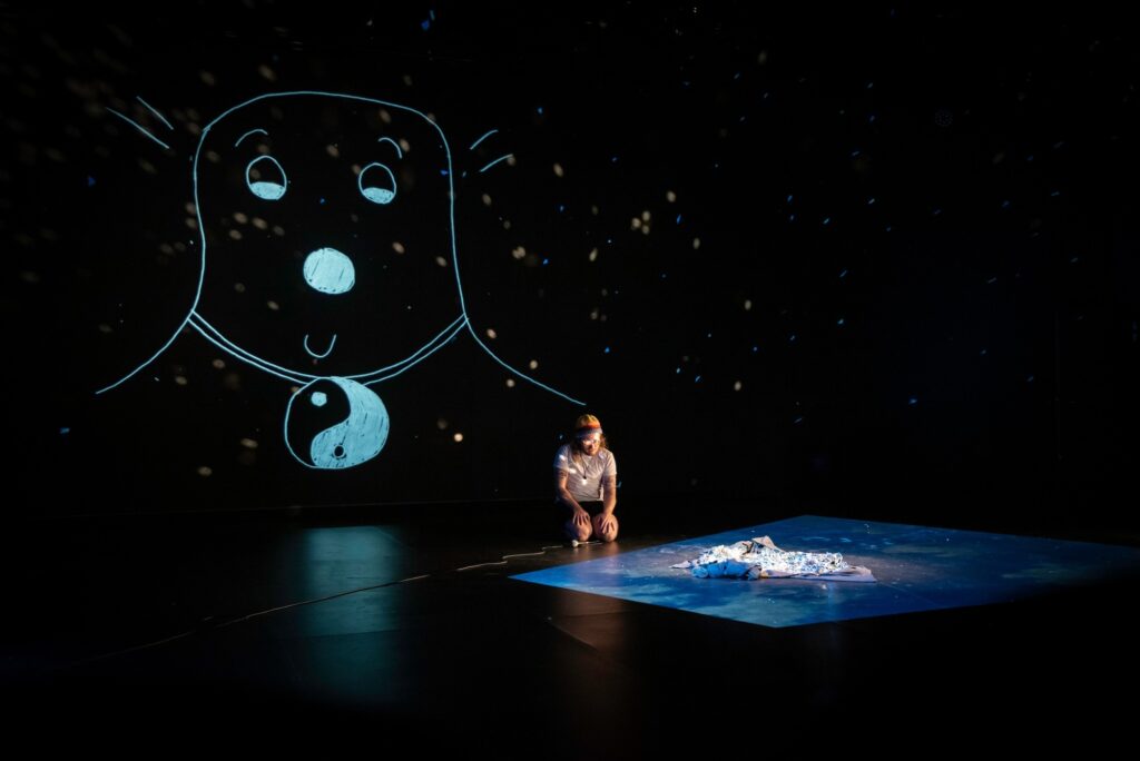 Person sits on black stage, which large drawing of figure with yinyang necklace behind them.