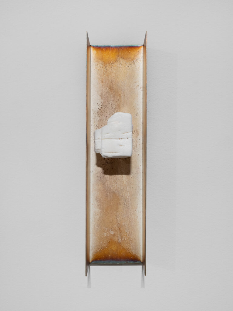 Cubic white stone-like object mounted on burnished bronze metal rectangle with sides bent forward