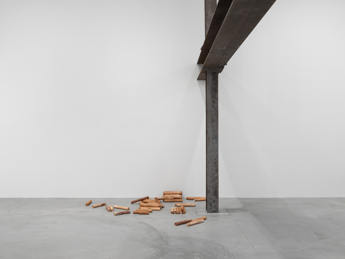 Large space with white walls, concrete floors, a large metal beam and post, and brown dowels spread and stacked at the base of the post