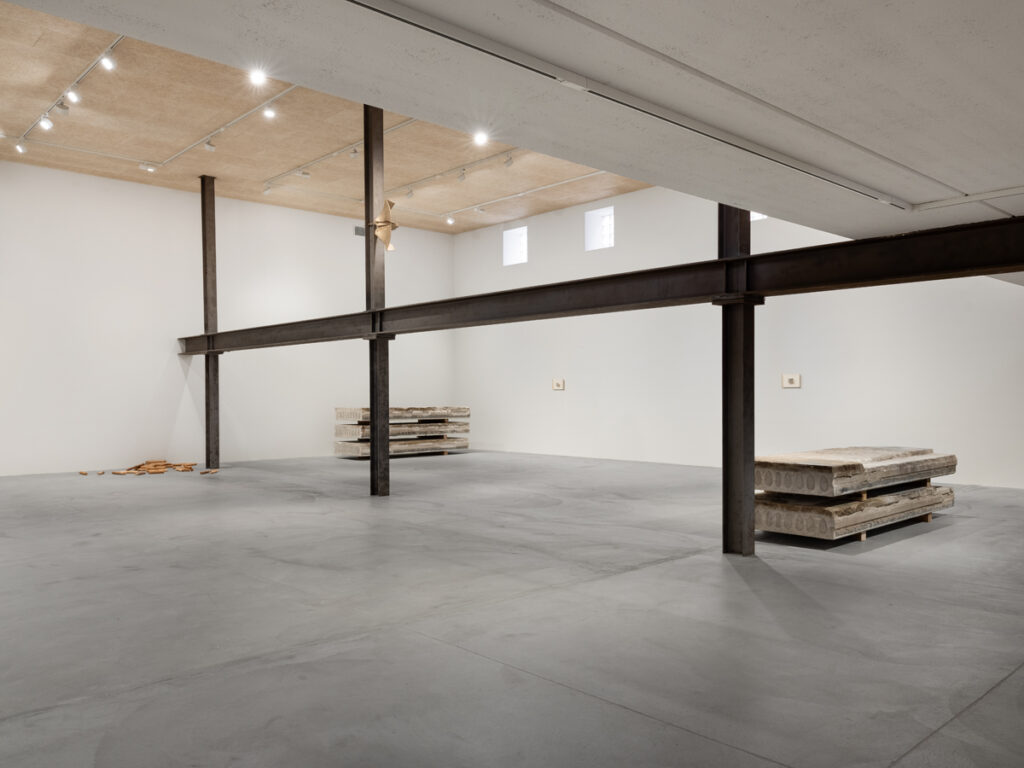 Large industrial space with metal beams, stacks of concrete rectangles on wooden feet, wooden dowels in far corner