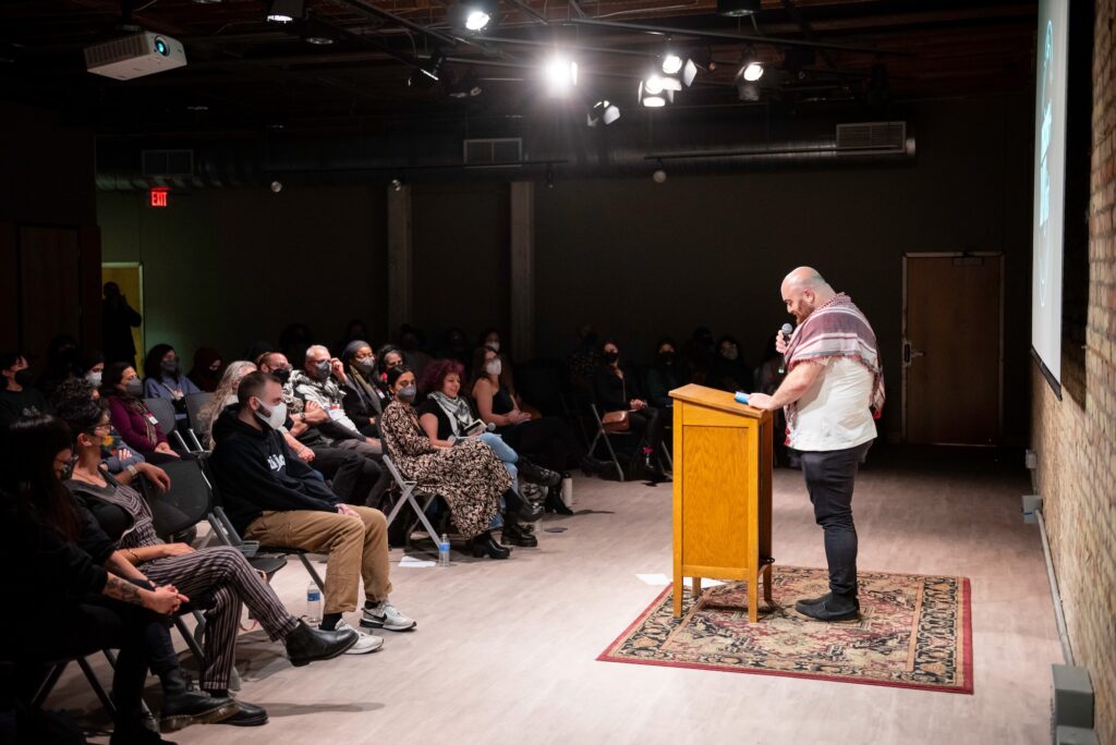 Person stands at podium with rug underneath, reading for an audience wearing masks