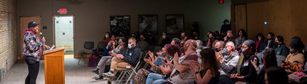 Person stands at podium smiling, reading for an audience clapping and wearing masks