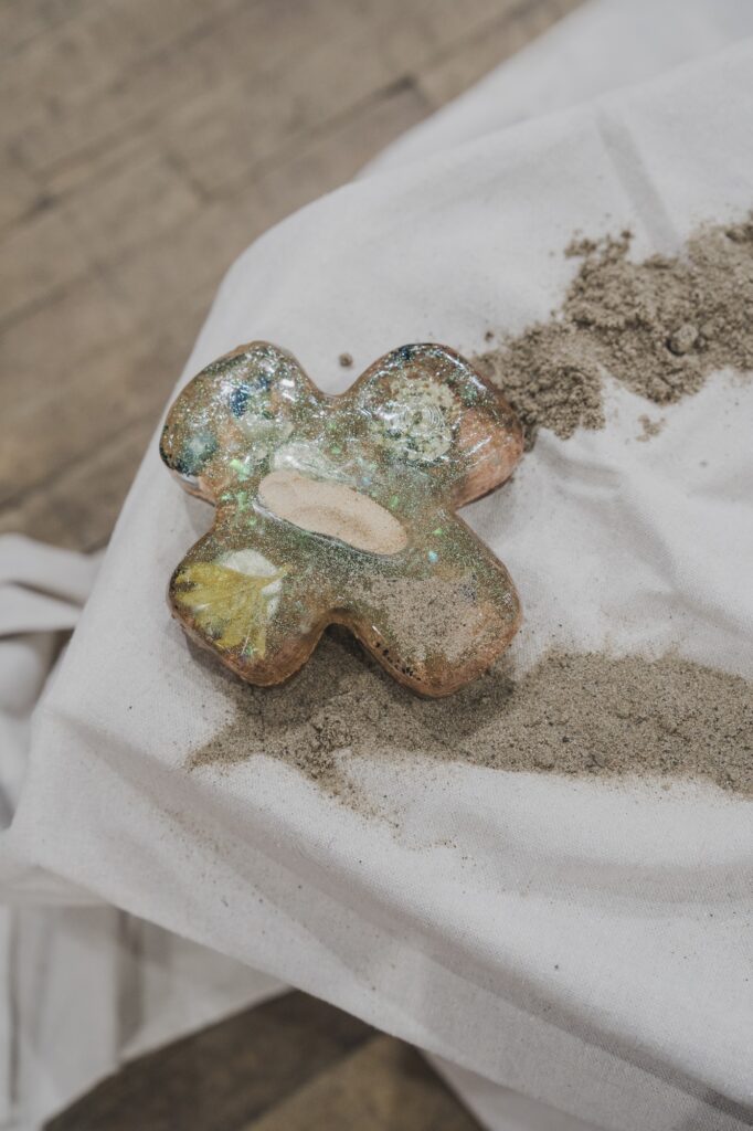 Resin object in a cross/clover shape flecked with shimmering dust and containing found objects.