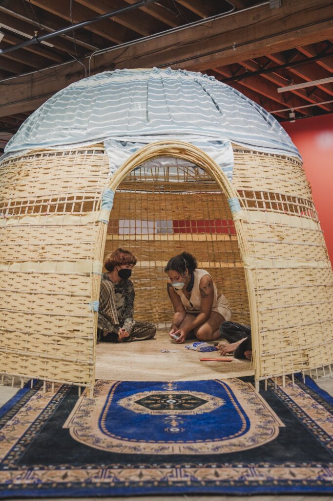 A dwelling made of bamboo and reeds with an oval opening and ornate blue carpet connecting inside and outside. Two masked people sit near each other inside the dwelling.