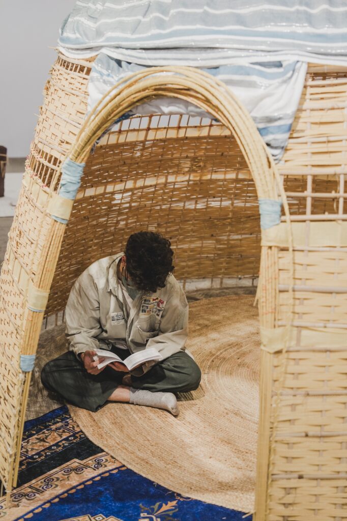 A dwelling made of bamboo and reeds with an oval opening and person reading inside.