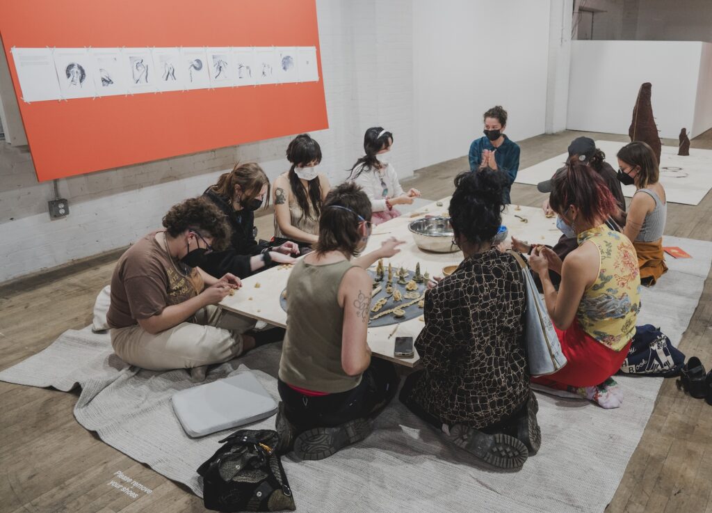 A circle of masked participants are seated on the floor, molding objects by hand.