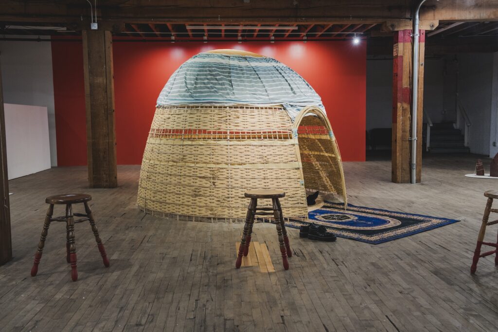 A dwelling made of bamboo and reeds with an oval opening and ornate blue carpet connecting inside and outside.