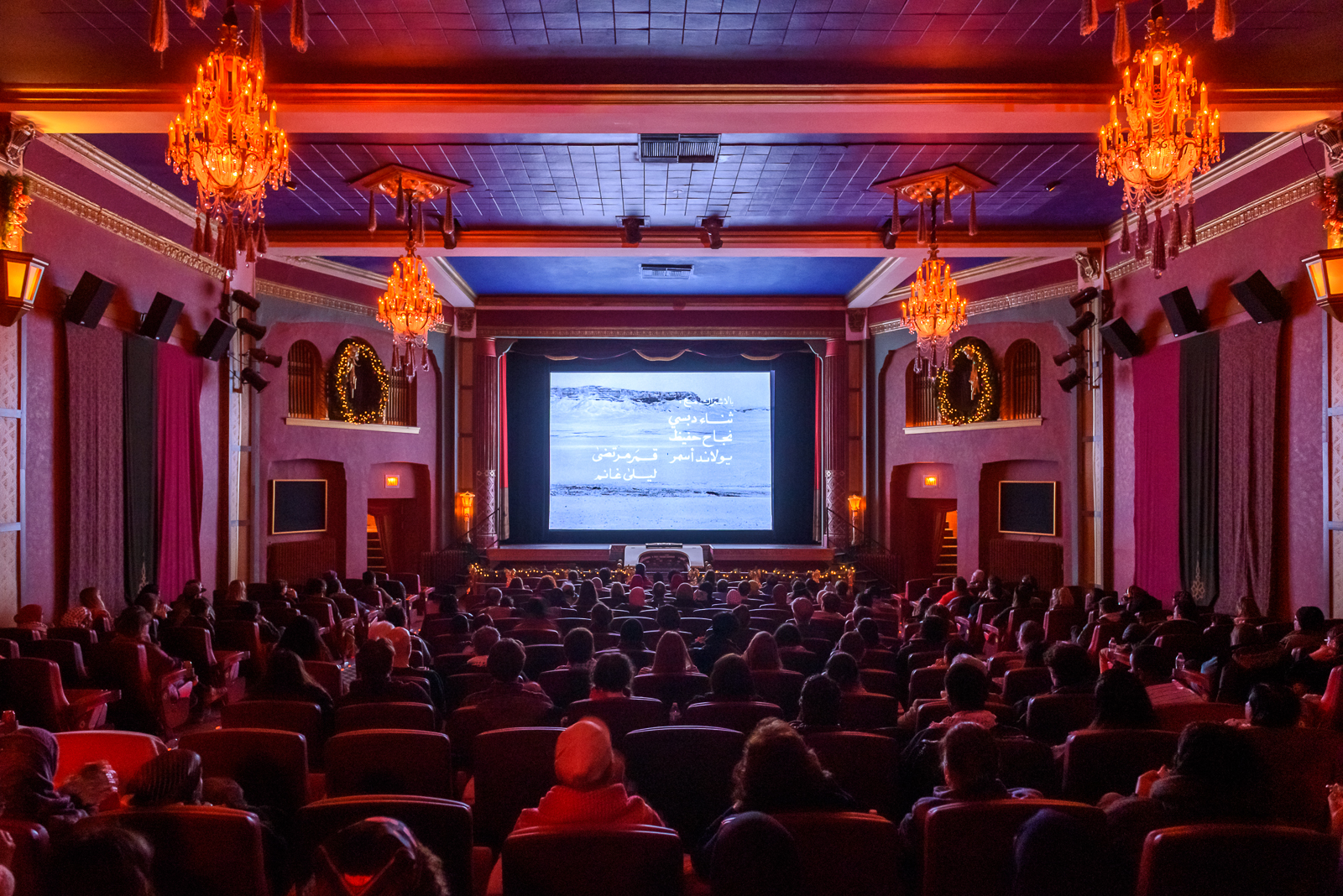 Photo from the back of a movie theater with ornate chandeliers, many people in seats and Arabic writing against a landscape on the screen.