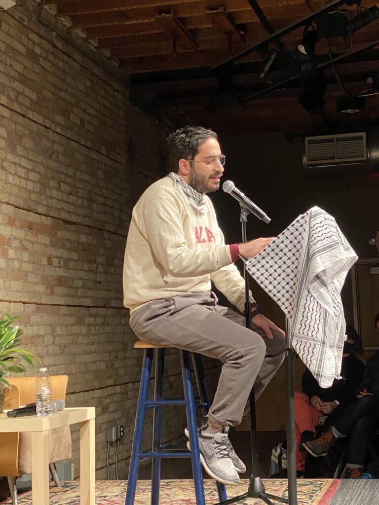 Person sits on stool reading into a microphone, with a Palestinian scarf covering the stand in front of them.