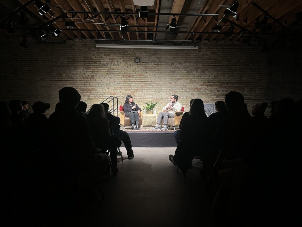 Two people sit on platform in chairs in front of an audience, with a brick wall behind them.