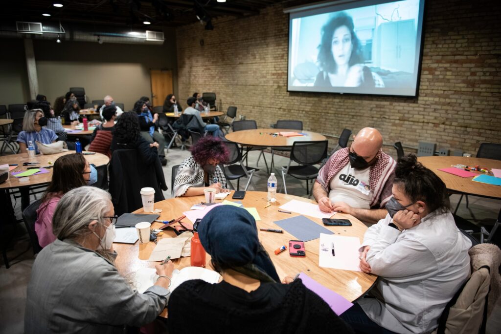 People sit around circular tables writing, a person projected on a screen behind them in mid-speech.