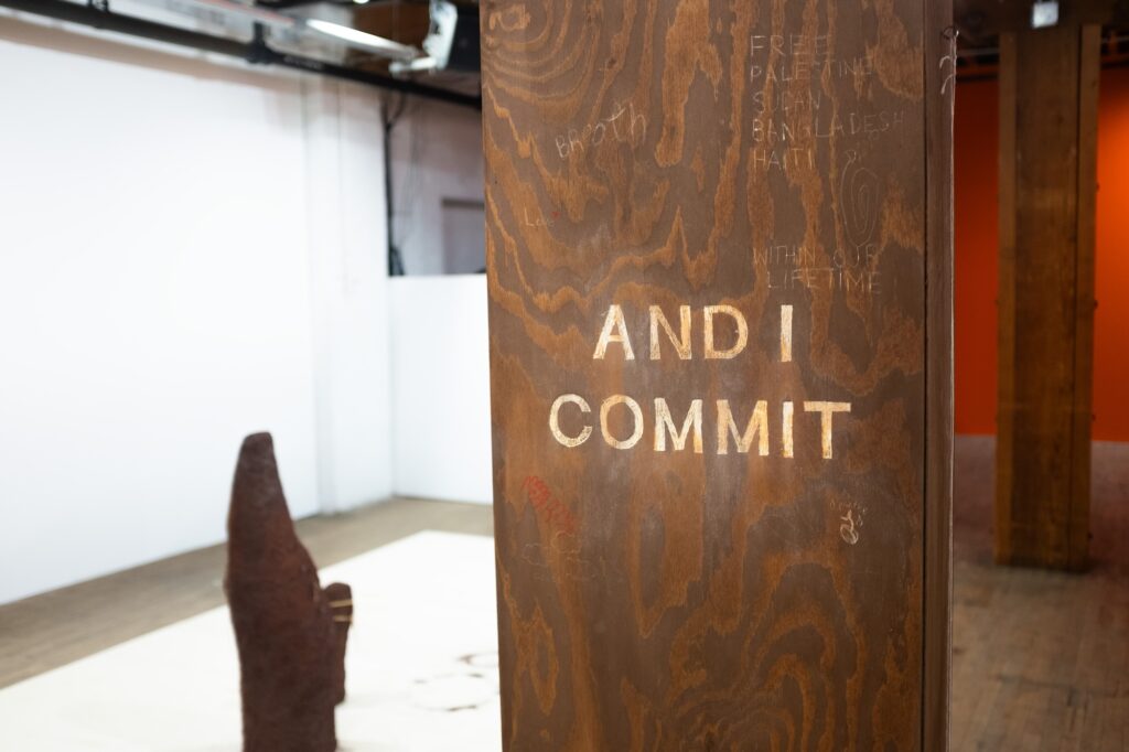 A wood panel with the words “AND I COMMIT” written on it. Engravings on the wood panel read “FREE PALESTINE SUDAN BANGLADESH HAITI IN OUR LIFETIME”